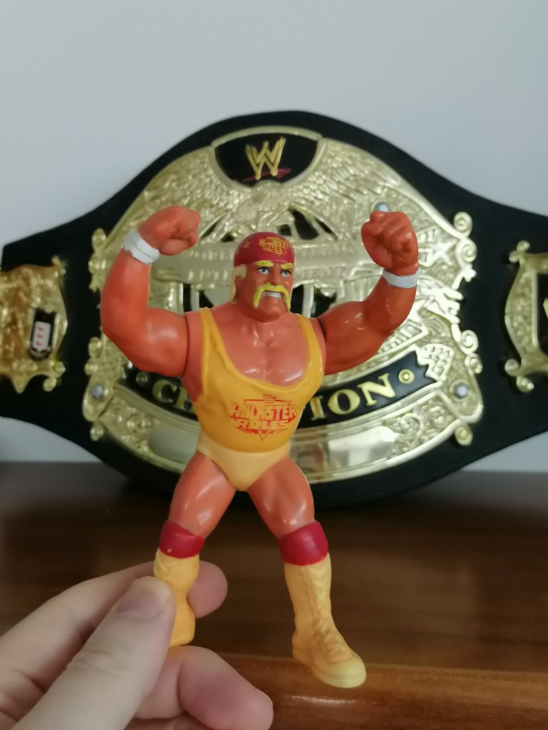 Vintage Hulk Hogan action figure in yellow gear with “HULKSTER RULES” shirt, posed in front of a golden WWE championship belt. Tribute by Leopoldo La Ciura.